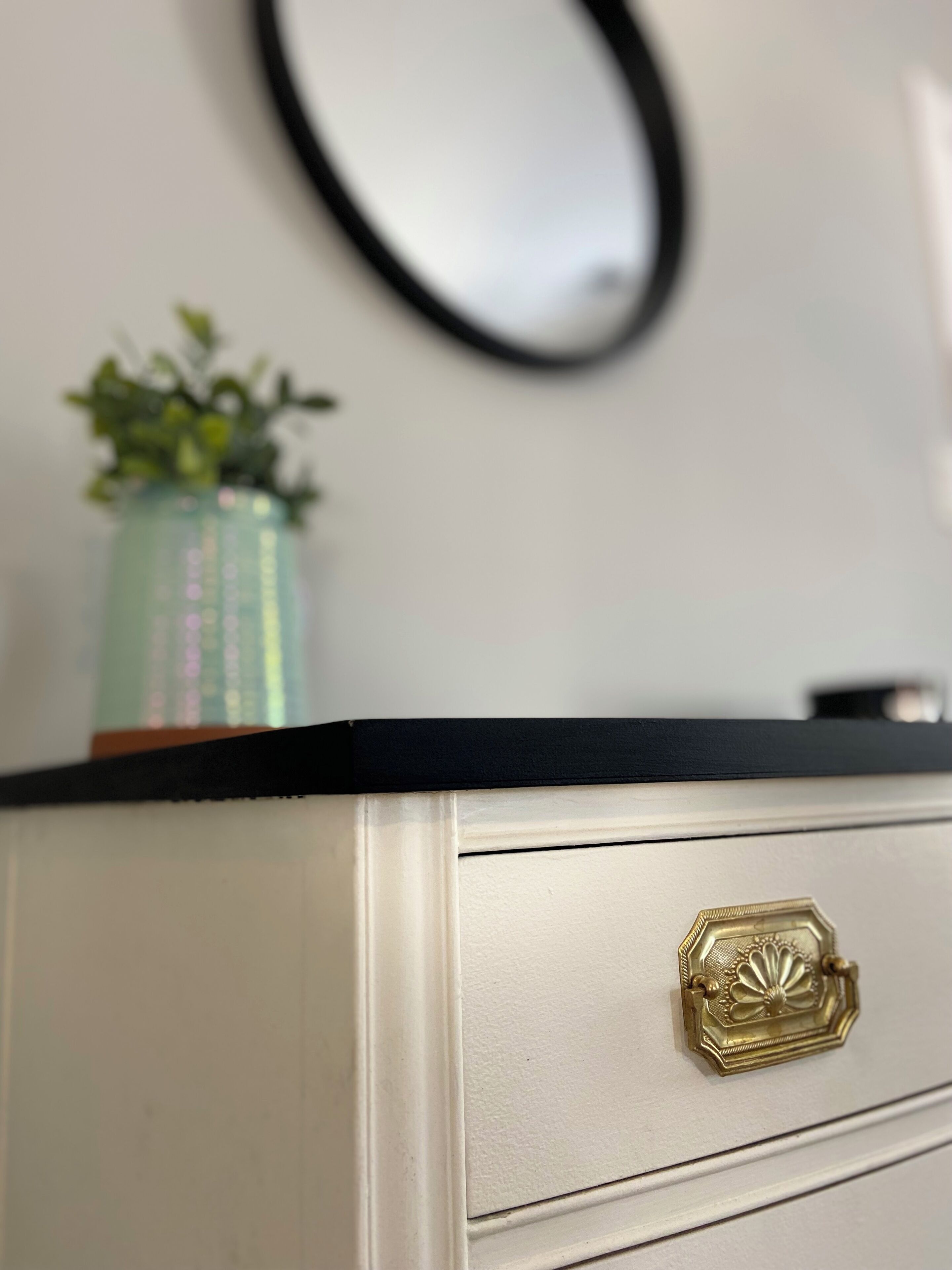 A white dresser with a black top, a gold handle, and a small green plant in a glass container.