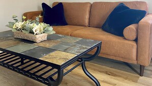 Un salon avec un canapé en cuir marron, une table basse en métal noir agrémentée d’un arrangement floral et un tableau abstrait aux couleurs vives accroché au mur.