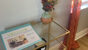 Un panneau d’accueil, un vase contenant des plantes séchées et un bol en cuivre, le tout posé sur une table à cadre doré, devant un mur orné d’un tableau.