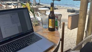 Una computadora portátil sobre una mesa de madera, con una botella de licor, un vaso y una planta. El fondo muestra una playa con sombrillas y palmeras.