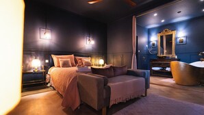 Ein Schlafzimmer mit einem Bett, zwei Nachttischen mit Lampen, einem großen Spiegel, einer Badewanne und einem Sessel.