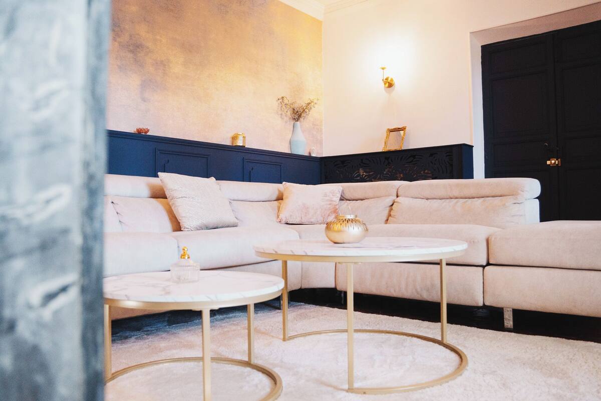 A modern living room with a beige sectional sofa, two gold-rimmed coffee tables, and a dark blue accent wall.