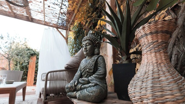 Eine Buddha-Statue auf einer hölzernen Bank in einem überdachten Außenbereich mit einem Korbwägelchen und einem Blumentopf in der Nähe.