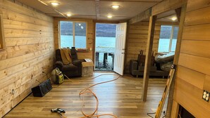 A wooden cabin with a large window overlooking a lake, a chair, a box, and construction tools on the floor.