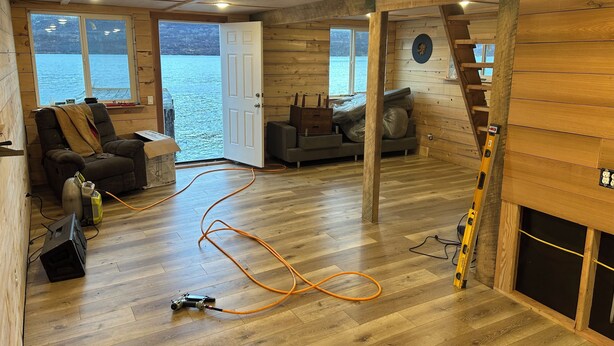 A cabin with a wooden interior, a large window overlooking a body of water, a sofa, a chair, and a ladder.