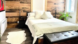A bedroom with a wooden wall panel, a bed with white bedding, a bedside lamp, a small table, a potted plant, and a white furry rug.