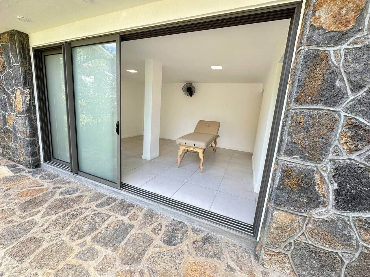 Un mur extérieur en pierre avec une porte-fenêtre coulissante donnant sur un espace intérieur où se trouve un fauteuil beige.