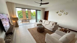 A modern living room with a sofa, coffee table, and TV. There is a balcony with a view of greenery.
