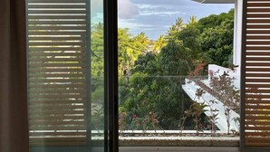 A modern balcony with a glass door, offering a view of lush greenery and a white building.