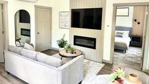 Un salon moderne avec un canapé gris, un téléviseur à écran plat fixé au mur, une petite table ronde agrémentée d’une plante et une cheminée.