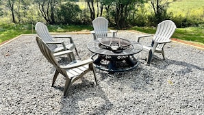 Un foyer est entouré de quatre chaises Adirondack, le tout installé sur un sol de gravier, dans un espace verdoyant avec des arbres en arrière-plan.