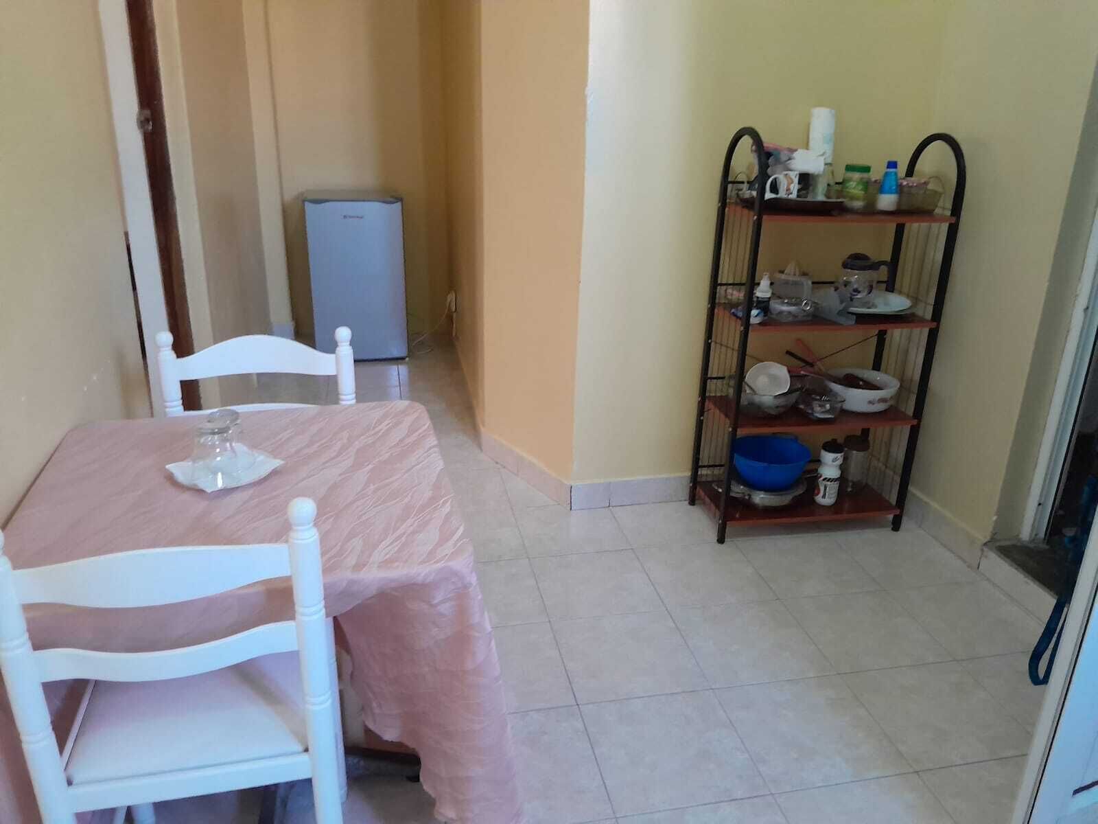 Un coin repas avec une table recouverte d’une nappe rose, des chaises blanches et une étagère en métal où sont disposés de la vaisselle et des bouteilles.