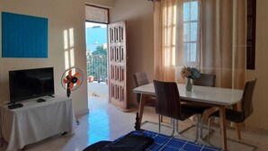 A room with a bed, a TV, a fan, a table with chairs, a window with curtains, and a door leading outside. - The Hidden One (Praia)