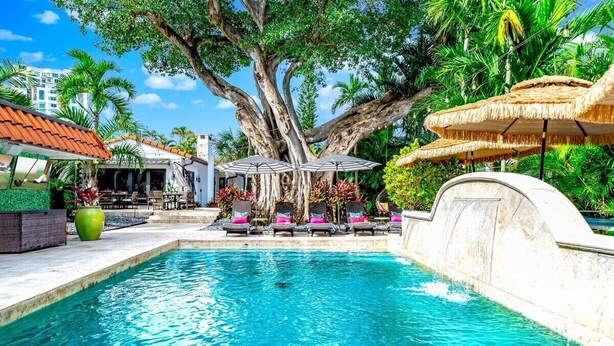 A swimming pool with lounge chairs and umbrellas, surrounded by tropical trees and a building with a red-tiled roof.