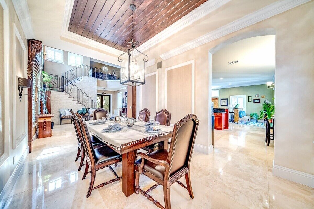 Un comedor espacioso con una gran mesa de madera, sillas elegantes y una araña. Una escalera conduce a un nivel superior.