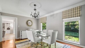 A dining area with a glass table, white chairs, a chandelier, and a view of greenery through large windows.