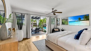 A bedroom with a bed, a ceiling fan, a large window with curtains, a view of a beach, and a painting of a beach scene. - New Waterfront Oasis, Minutes to Beach, Eats, Shops (Fort Lauderdale)