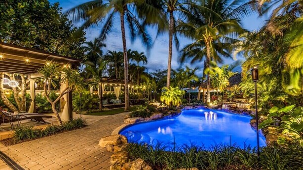 A pool area with a curved edge, surrounded by rocks and tropical plants, and a covered seating area with string lights. - New Waterfront Oasis, Minutes to Beach, Eats, Shops (Fort Lauderdale)