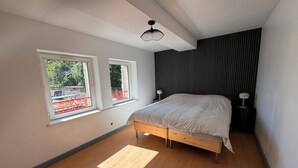 Un dormitorio con cama, dos ventanas, una lámpara de techo y una mesita de noche.