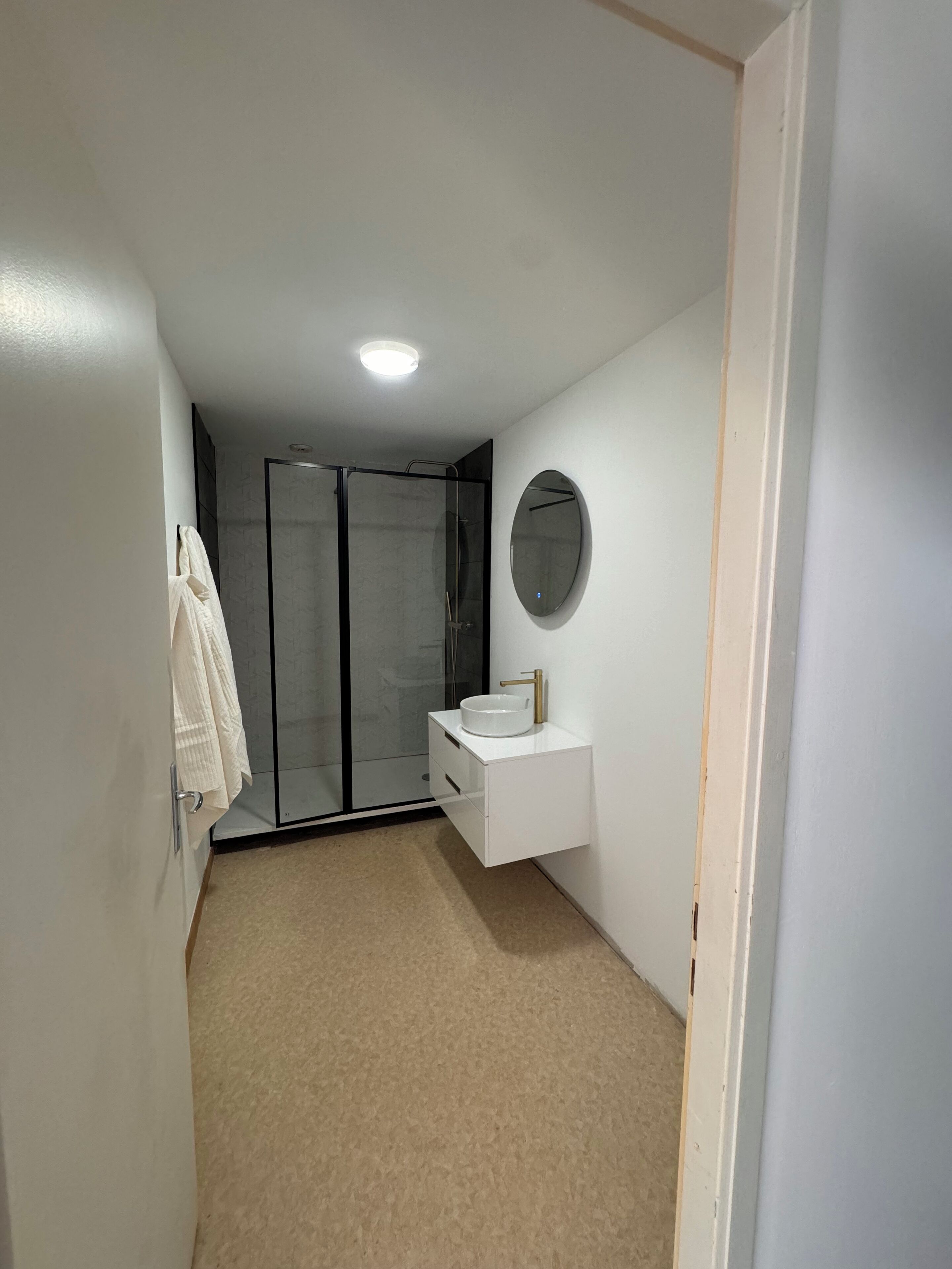 Une salle de bain avec une douche à parois de verre, un lavabo blanc avec un robinet doré et un miroir rond accroché au mur.