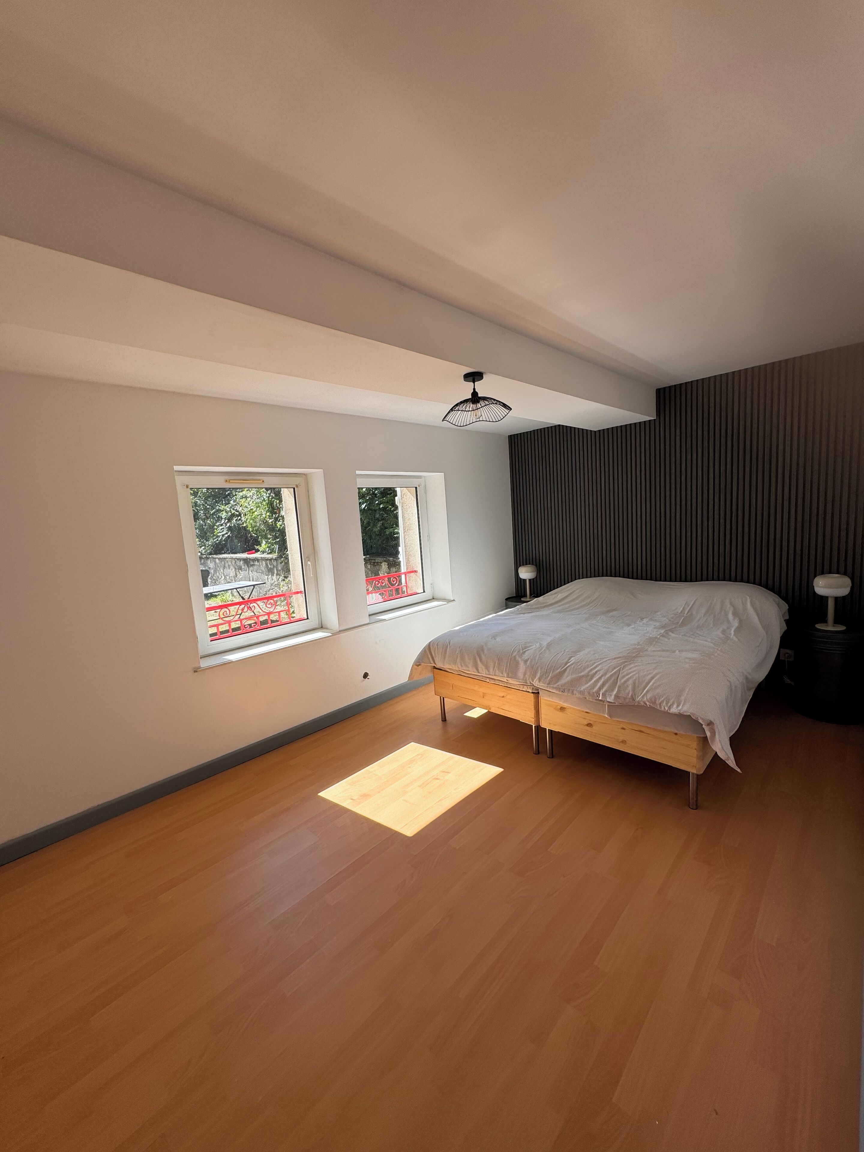 Un dormitorio con una cama, dos ventanas, una lámpara en el techo y suelo de madera.