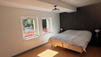 Un dormitorio con una cama, dos ventanas, una lámpara en el techo y suelo de madera.