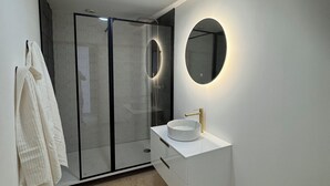 Un baño moderno con ducha envuelta en vidrio, tocador blanco con grifo dorado y espejo redondo con luz.