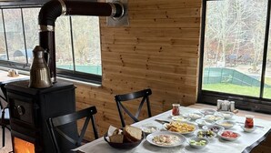 A cozy cabin interior with a wood-burning stove, a dining table set with various dishes, and a view of a fenced yard through a large window. - Abant Barış Pension (Bolu)