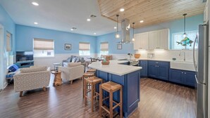 A modern kitchen with blue cabinets, a central island, wooden stools, and a dining area with a sofa and chairs. - Luxe Coastal Stay w/ Private Pool, Walk to Beach Leave of Absence by Avantstay (Port Aransas)