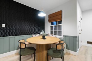 Ein Essbereich mit einem runden Holztisch, vier Stühlen, einer hängenden Leuchte, einem Fenster mit brauner Jalousie und einer Wand mit Muster.