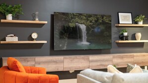 A modern living room with a large flat-screen TV mounted on the wall, wooden shelves with decorative items, and two sofas.