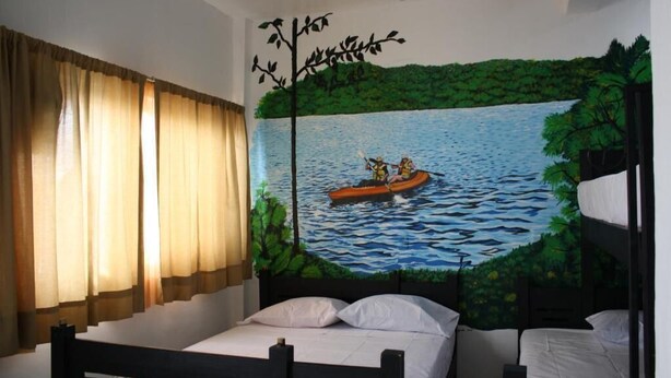 A bedroom with a mural of a river scene, a bed with white bedding, and a window with curtains. - HOTEL CALLE 8 (Tuxtla Gutierrez)