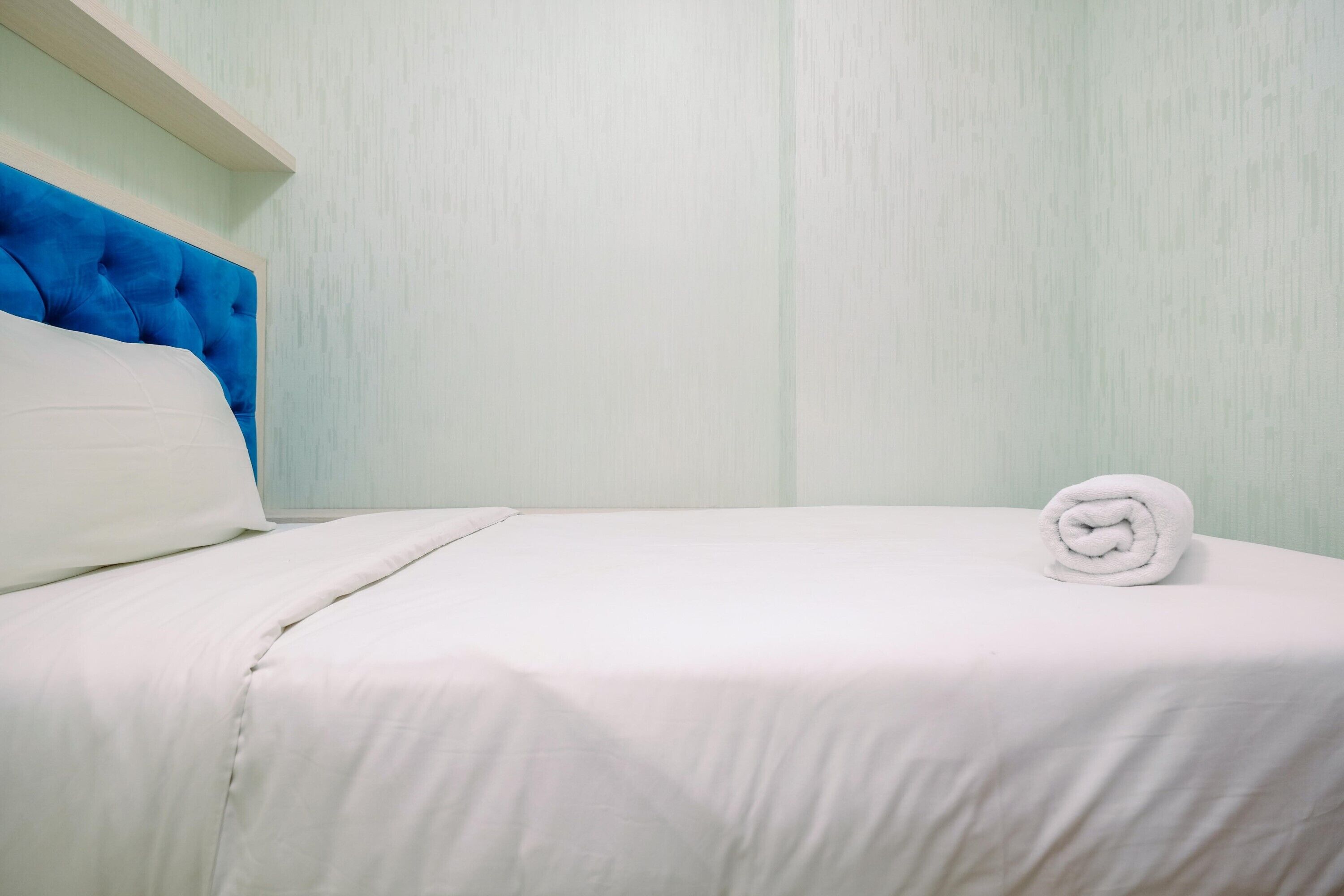 A neatly made bed with white linens and a rolled towel on the side, a blue headboard, and a light-colored wall.