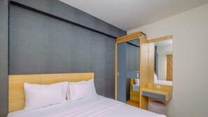 A hotel room with a bed, a wooden headboard, a gray wall, a wooden side table, a mirror, and a bathroom with a sink and towel. - Best Choice and Tidy 2BR Podomoro Golf View Apartment (Gunung Putri)
