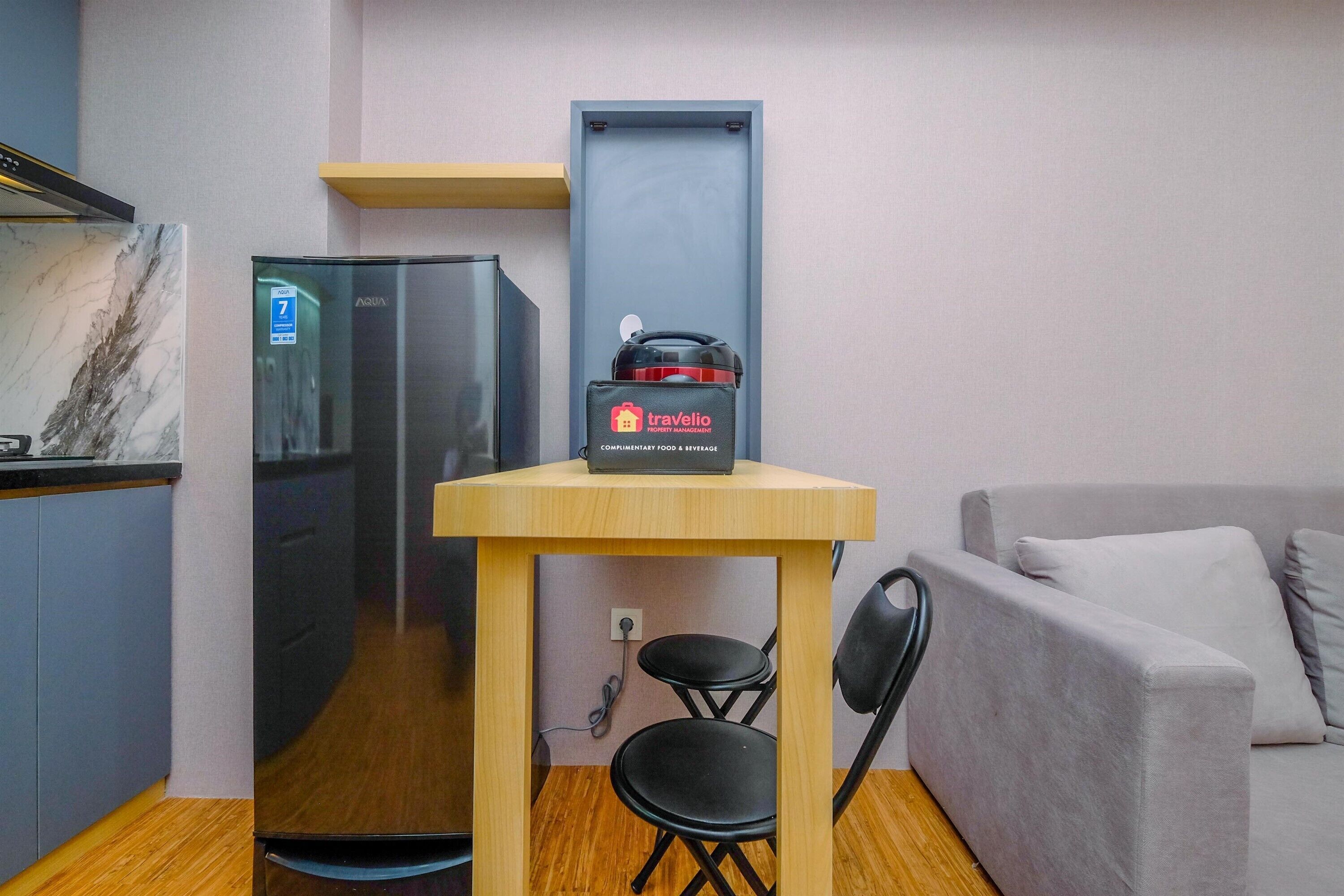 A compact kitchenette with a refrigerator, a travel bag on a small table, and a sofa in the background.