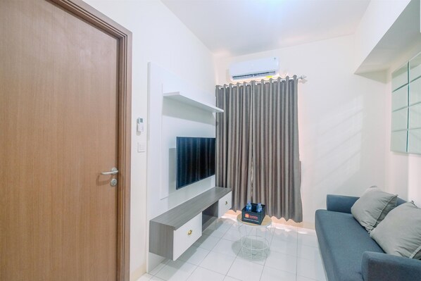 A modern living room with a flat-screen TV, a grey sofa, a coffee table, and a grey curtain. - Cozy Living and Relaxing Podomoro Golf View 2BR Apartment (Gunung Putri)