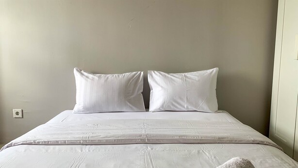 A bed with white bedding and pillows, a plain wall, and a door on the right. - Cozy Studio at Belmont Residence Puri Apartment (Jakarta)