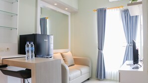 A modern hotel room with a sofa, a desk with a computer, and a TV. There are two water bottles on the desk. - Spacious and Good 2BR at Podomoro Golf View Apartment (Gunung Putri)