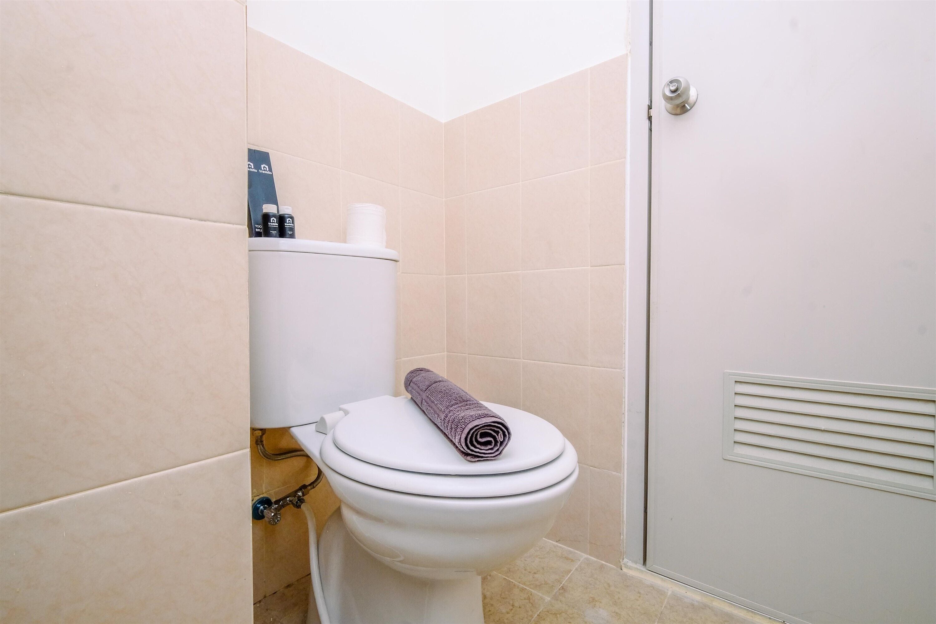 Une salle de bain avec des toilettes, des carreaux de couleur beige, une serviette posée sur le couvercle des toilettes et une porte avec une poignée.