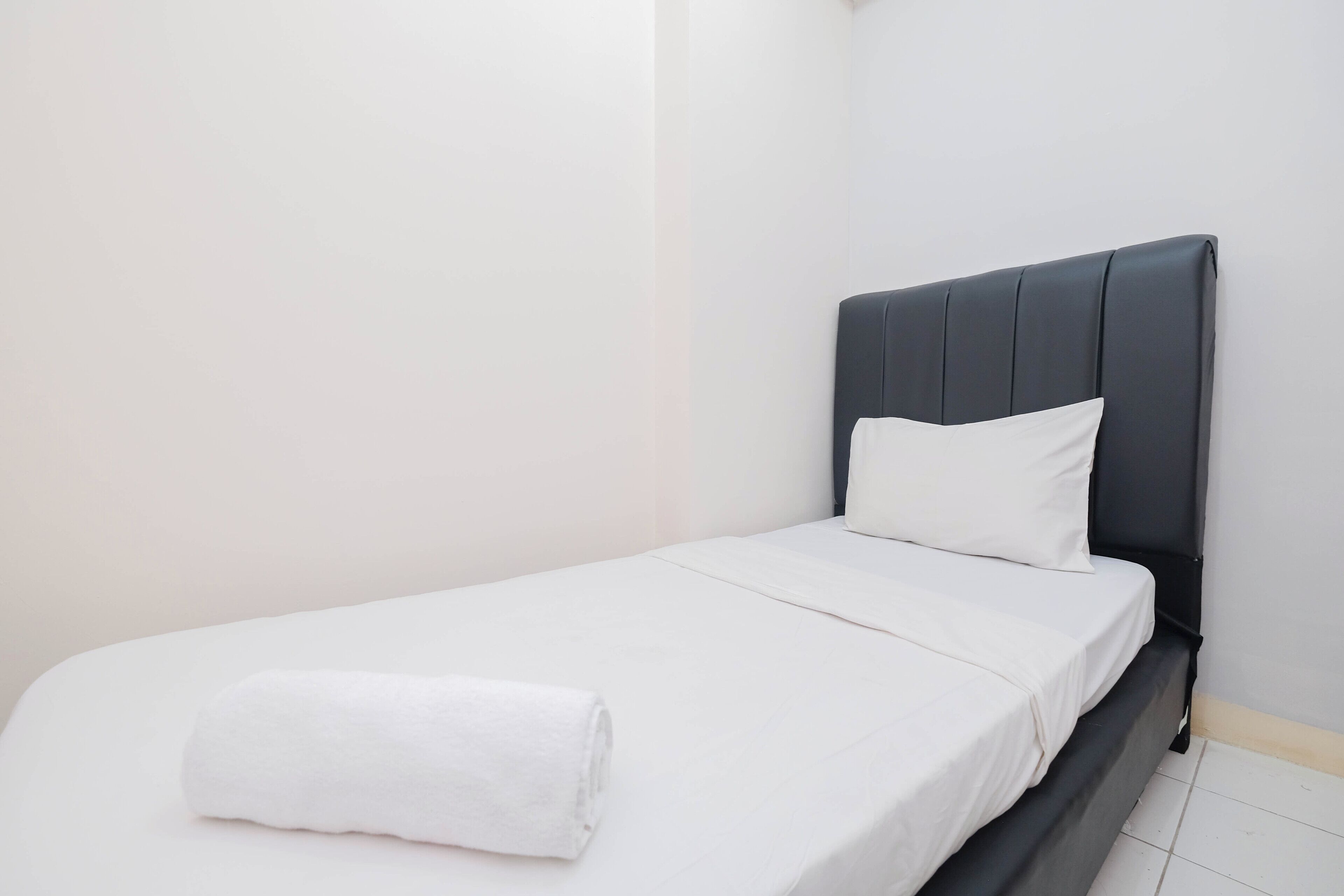Ein Einzelbett mit dunklem Kopfteil, weißer Bettwäsche und einem aufgerollten weißen Handtuch auf dem Bett.
