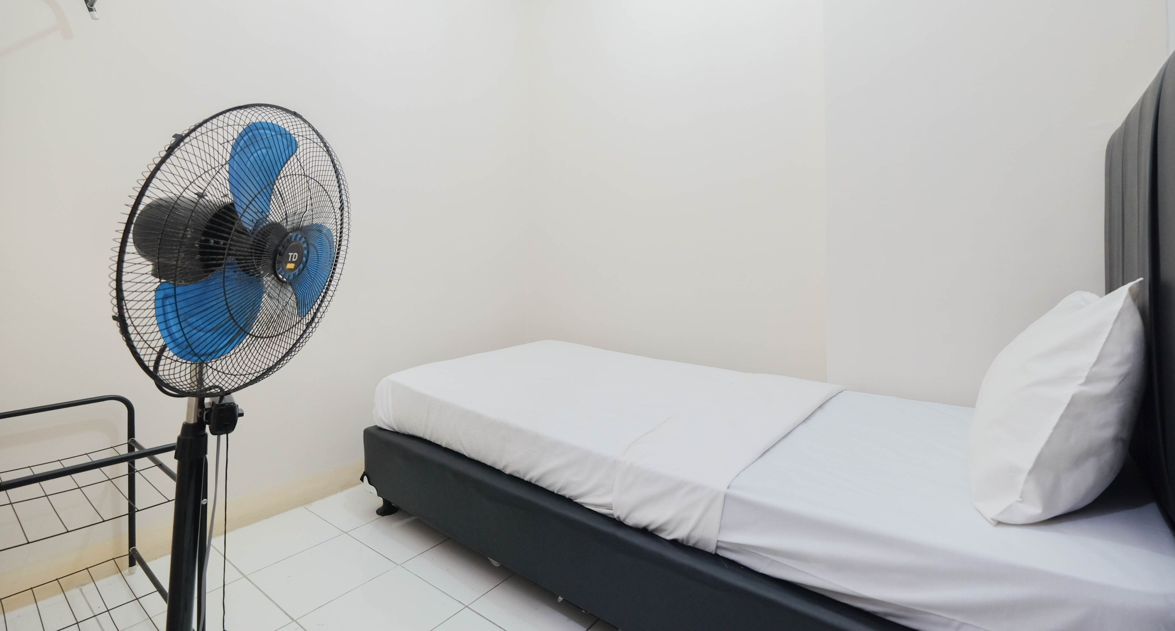 A small, single-bed room with a standing fan, a metal rack, and a plain white wall.