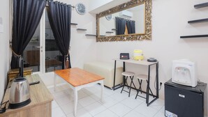 A compact room with a wooden table, a white sofa, a small stool, a mirror, a clock, a water dispenser, and a kettle.
