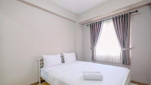 A neatly made bed with white linens and a folded towel, a window with curtains, and a wooden floor.