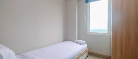 A small, neatly made bed in a room with a window, a wooden wardrobe, and a light-colored floor.