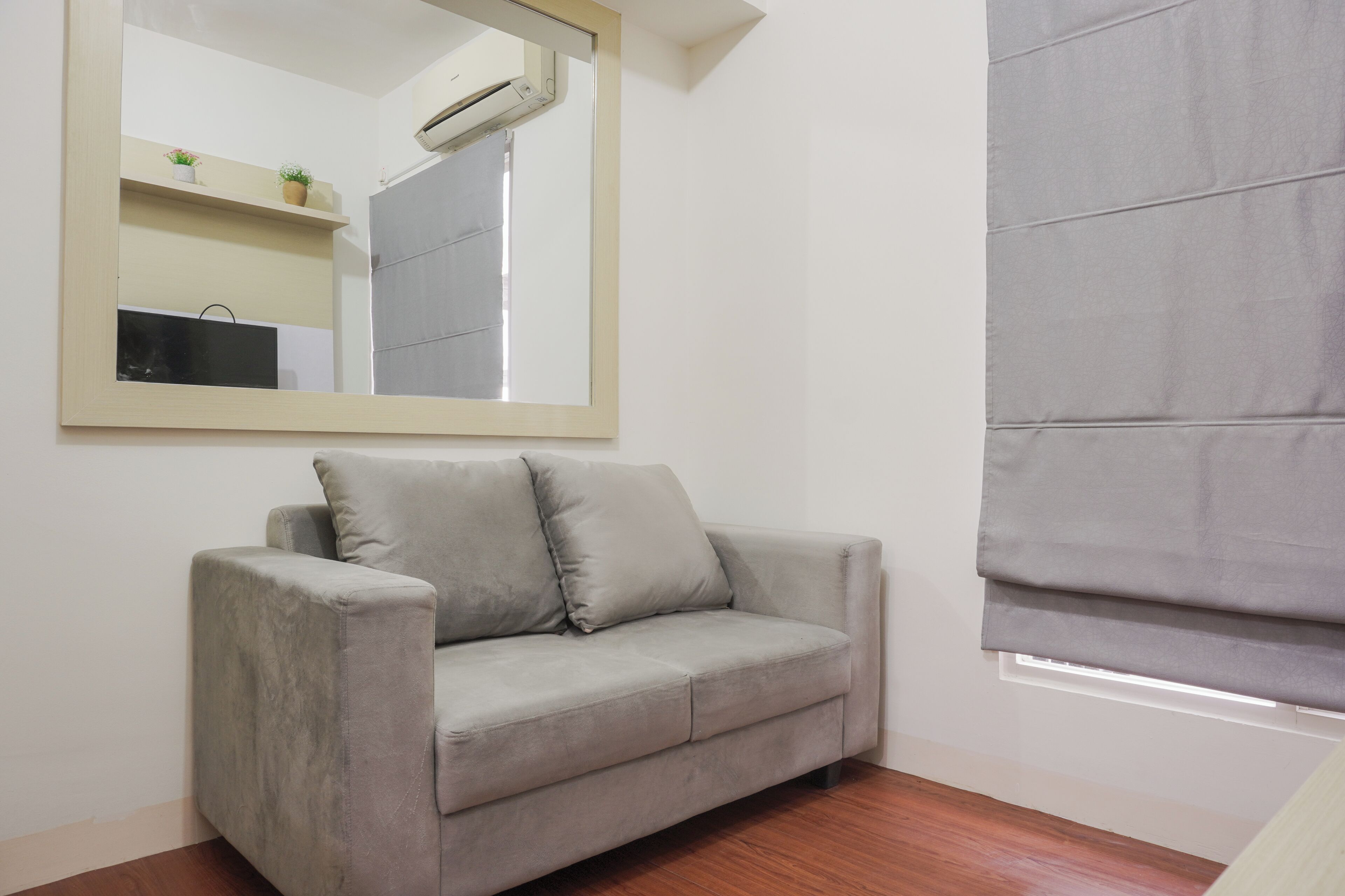 A room with a grey sofa, a large mirror, a window with grey blinds, and a wooden floor.