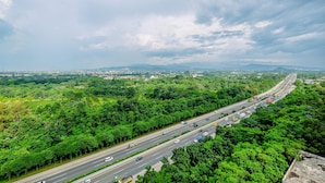 A highway with multiple lanes, surrounded by dense green vegetation and distant mountains under a cloudy sky.