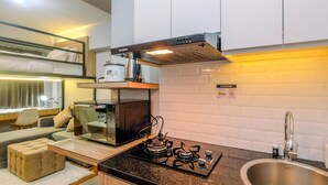 A compact kitchen with a gas stove, a microwave, and a sink. There is a sofa and a small ottoman in the living area. - Enjoy and New Furnished Studio at Podomoro Golf View Apartment (Gunung Putri)