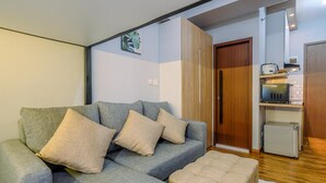 A living room with a grey sofa and ottoman, a wooden door, and a kitchen area with a small fridge and a toaster. - Enjoy and New Furnished Studio at Podomoro Golf View Apartment (Gunung Putri)