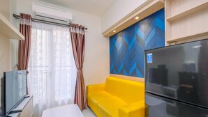 A modern living room with a yellow sofa, a flat-screen TV, a refrigerator, and a window with curtains. - Simply Design 2BR at Podomoro Golf View Apartment (Gunung Putri)