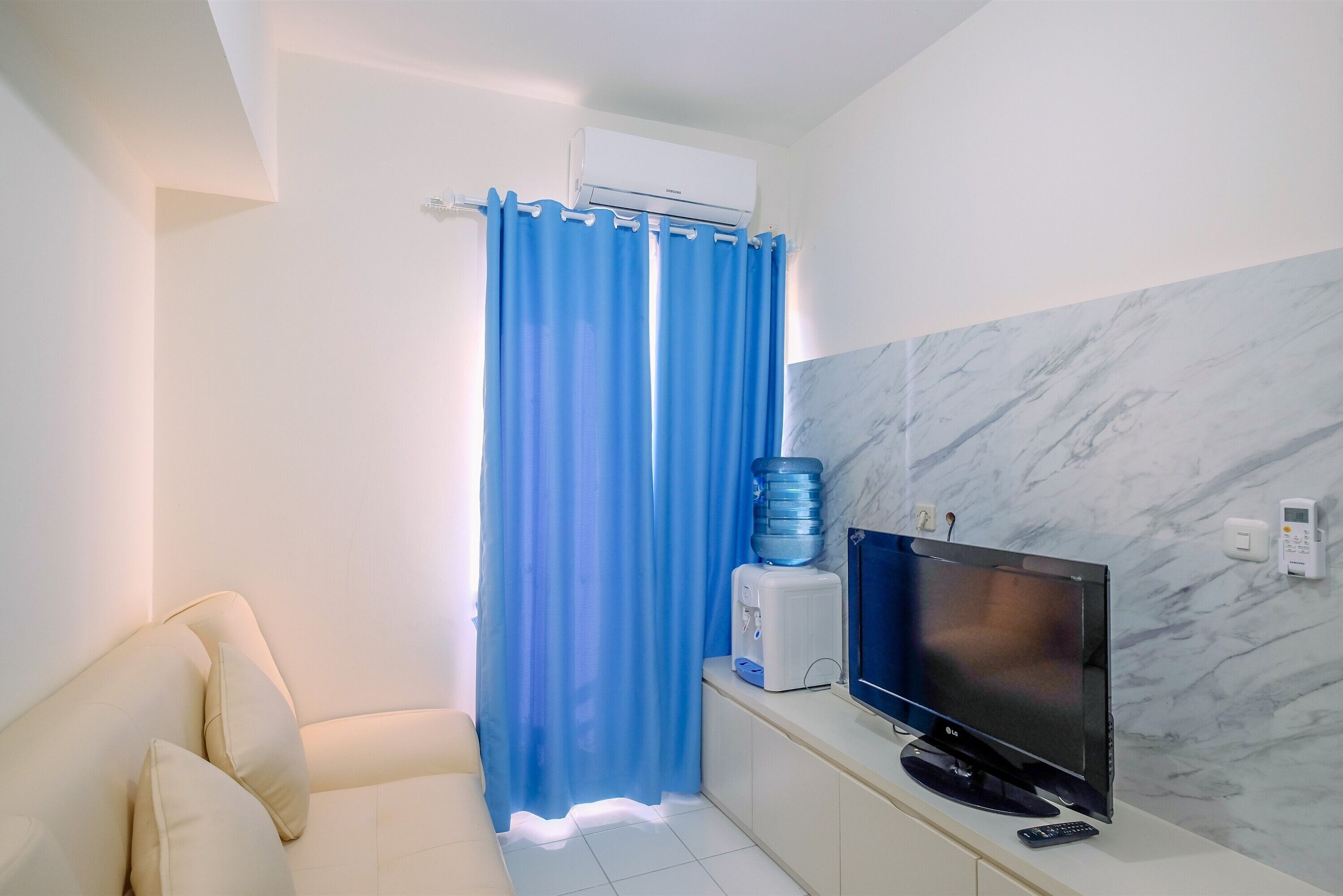 A room with a sofa, a flat-screen TV, a water dispenser, and a marble-patterned wall.